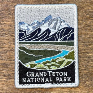 Può includere: Toppa ricamata raffigurante una catena montuosa con cime innevate, un fiume e la scritta "GRAND TETON NATIONAL PARK". La toppa ha un bordo grigio e una base nera con testo bianco.