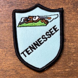 May include: A shield-shaped embroidered patch with a light blue background and a black stitched border. It features a map of Tennessee in white with a brown and green graphic inside, and the word TENNESSEE stitched in black capital letters.