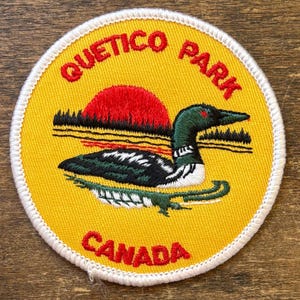 May include: A round yellow patch with the words "QUETICO PARK CANADA" in red. The patch features a stylized illustration of a loon with a red sunset and a green and white loon. The patch has a white border.