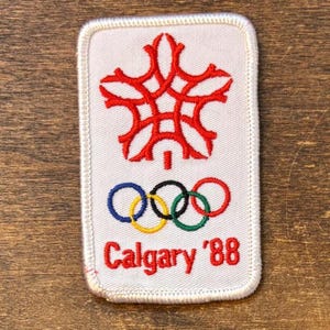 Puede incluir: Un parche rectangular con fondo blanco presenta un emblema rojo en la parte superior, los anillos olímpicos y el texto "Calgary '88" en rojo. El parche tiene un borde blanco y es probablemente un recuerdo del evento.