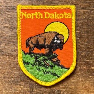 May include: Embroidered patch with a bison on a grassy hill, a yellow sun, and an orange background. The patch has a yellow border and the text "North Dakota". The patch is likely a souvenir or emblem.