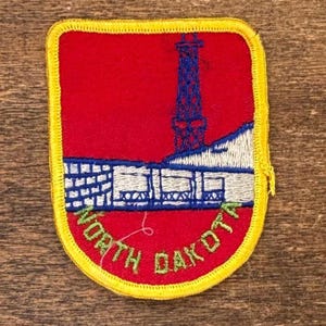 May include: Embroidered patch with a red backdrop, depicting a blue and white industrial structure. The patch is edged with yellow and has the words "NORTH DAKOTA" in green.