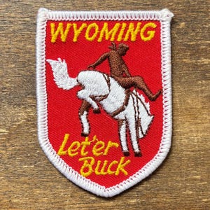 Puede incluir: Parche bordado con fondo rojo y borde blanco. El parche presenta un bronco con un jinete y las palabras "WYOMING" y "Let'er Buck" en amarillo.