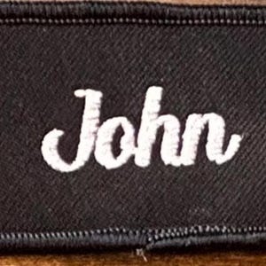 以下が含まれることがあります： 白い筆記体で「John」と書かれた黒い刺繍パッチ。 パッチは、ステッチされたエッジを持つ黒い境界線を持っています。
