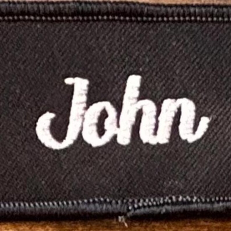 以下が含まれることがあります： 白い筆記体で「John」と書かれた黒い刺繍パッチ。 パッチは、ステッチされたエッジを持つ黒い境界線を持っています。