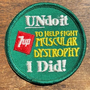 May include: A round green embroidered patch featuring the white text UNdoit, a red square with the white 7up logo, yellow text reading TO HELP FIGHT MUSCULAR DYSTROPHY, and the white text I Did! at the bottom with a dark green stitched border.