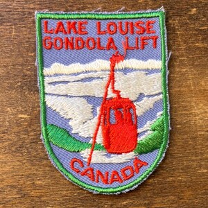 May include: Embroidered patch featuring the text "Lake Louise Gondola Lift Canada." The patch has a light blue background with a red gondola and white clouds. Green and red stitching outlines the patch.