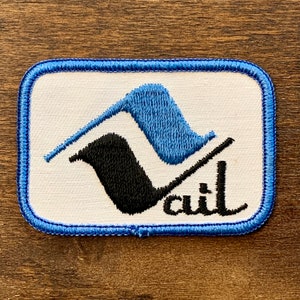 May include: Rectangular patch with a white background, blue border, and embroidered design. The design features a blue and black stylized mountain shape with the word "Vail" in black script.