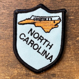 May include: A shield-shaped embroidered patch with a light blue background and a thick black border. It features an orange map of North Carolina with a small black building icon and the black embroidered text NORTH CAROLINA below the map.