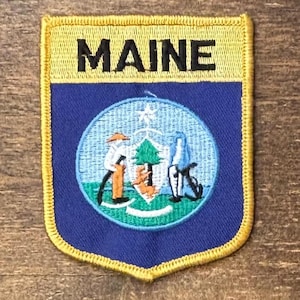 May include: A shield-shaped embroidered patch with a yellow border. The top section is yellow with the word MAINE in black text. The lower section is blue and features a circular emblem depicting a central pine tree, a farmer, and a sailor.