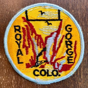 Puede incluir: Parche redondo con fondo amarillo y borde azul claro. El parche presenta las palabras "ROYAL GORGE COLO." y "GORGE" en texto negro. Un diseño rojo y blanco está en el centro, con una línea negra y dos pájaros arriba.
