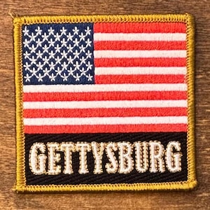 May include: A square embroidered patch featuring the United States flag in the upper section with a blue field of white stars and red and white stripes. The lower section is a black rectangular field with the word GETTYSBURG in white text.