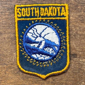 May include: A navy blue and gold embroidered patch with the text "SOUTH DAKOTA". The patch is shield-shaped and features a blue and white circular design in the centre, depicting a landscape. The patch is edged with gold thread.