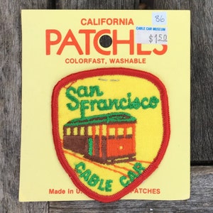 Puede incluir: Un parche bordado amarillo con un borde rojo que presenta un tranvía delante del texto "San Francisco" y las palabras "Cable Car" debajo. El parche está en una tarjeta amarilla con el texto "California Patches Colorfast, Washable Cable Car Museum $1.50" impreso en él.