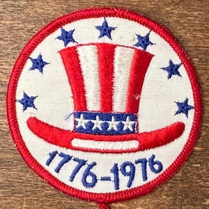 Peut inclure: Écusson circulaire avec une bordure rouge et un fond blanc. L'écusson présente un motif de chapeau de l'Oncle Sam rouge, blanc et bleu, entouré d'étoiles bleues. Le texte "1776-1976" est écrit en bleu.