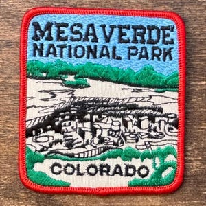 May include: Embroidered patch featuring the text "MESA VERDE NATIONAL PARK COLORADO." The patch has a blue sky, green trees, and a detailed illustration of the park's landscape. The patch is bordered with a red stitched edge.