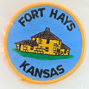 May include: A circular patch with a light blue background and a gold border. The patch features a yellow building with a brown roof and the words "FORT HAYS" and "KANSAS" in black lettering.
