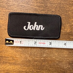 以下が含まれることがあります： 黒い長方形のパッチで、白い筆記体で「John」という名前が刺繍されています。パッチの長さは約7.6cmで、Craftsmanの巻尺で測定されています。