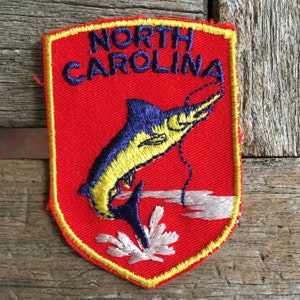 May include: Embroidered patch featuring a marlin leaping from the water. The patch is red with a yellow border and the words "NORTH CAROLINA" in blue. The marlin is blue and yellow, with white accents.