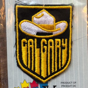 Könnte beinhalten: Gestickter Aufnäher mit einem Cowboyhut-Design über dem Wort "CALGARY" in Gelb, umrandet von Schwarz. Der Aufnäher hat einen schwarzen Hintergrund und einen gelben Rand. Der Hut ist weiß mit einer gelben Krempe. Ein Souvenirartikel.