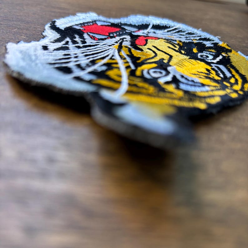 May include: An embroidered patch featuring a stylized tiger head design. The tiger is depicted with vibrant yellow fur, black stripes, white whiskers, and a bright red accent near the top. The patch has a white border and a textured surface.