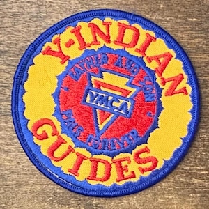 May include: A circular embroidered patch with a blue border. The outer ring is yellow with red text reading Y-INDIAN GUIDES. The centre is red with a blue triangle containing the text YMCA and the words FATHER AND SON PALS FOREVER.