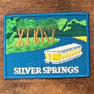 May include: Embroidered patch featuring a river scene with a boat, trees, and the text "Silver Springs" in white. The patch has a blue border and depicts a yellow and white boat on a blue river, with green trees and brown tree trunks.