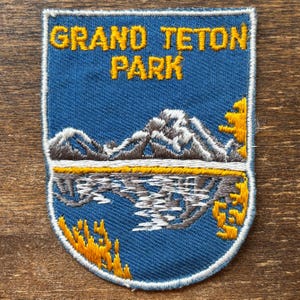 May include: A shield-shaped embroidered patch with a blue background featuring the yellow text GRAND TETON PARK. The design depicts a mountain range with white snow-capped peaks, a reflective body of water, and yellow foliage on the sides.