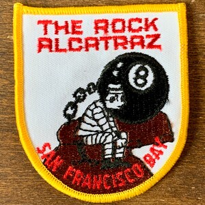 以下が含まれることがあります： 黄色の縁取りのある白い刺繍パッチ。パッチには、縞模様の服を着た人が黒い8ボールを持っているという漫画のイラストが描かれています。「THE ROCK ALCATRAZ」という文字がイラストの上に赤色で刺繍されています。「SAN FRANCISCO BAY」という文字がイラストの下に赤色で刺繍されています。