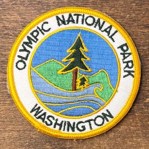 Könnte beinhalten: Ein kreisförmiger, bestickter Aufnäher mit blauem Himmel, grünen Bergen und einer dunkelgrünen Tanne in der Mitte. Der weiße Rand enthält den schwarzen Text OLYMPIC NATIONAL PARK WASHINGTON, umrandet von einer leuchtend gelben Kante.