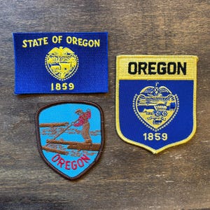 Puede incluir: Tres parches bordados con el estado de Oregón. Uno es rectangular con "STATE OF OREGON" y "1859". Otro tiene forma de escudo con "OREGON" y "1859". El tercero tiene forma de escudo con un leñador y la palabra "OREGON".