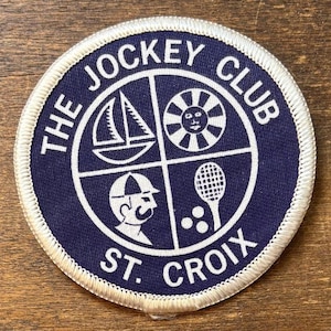 May include: A circular patch with a navy blue background and white embroidered details. The text "THE JOCKEY CLUB ST. CROIX" surrounds a segmented design with a sailboat, sun, tennis racket, and a person wearing a cap.