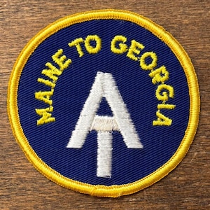 May include: A circular patch with a blue background and a yellow border. The text "MAINE TO GEORGIA" arches around the top in yellow. A white "A" with an upward arrow is in the centre. The patch is likely for a hiking trail.
