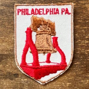 May include: Embroidered patch featuring the Liberty Bell in brown and red, with the text "PHILADELPHIA PA." in red. The patch has a white background and a shield shape, with a brown border.