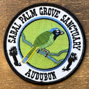 May include: A circular embroidered patch featuring a green bird perched on a blue branch against a light green background. The white border contains the black text "SABAL PALM GROVE SANCTUARY" and "AUDUBON" with two small black palm tree silhouettes.