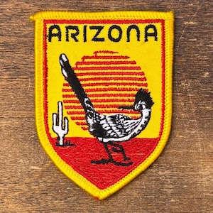 May include: Embroidered patch with a yellow border and a red shield shape. The patch features the word "ARIZONA" in black, a roadrunner, a setting sun, and a cactus. The roadrunner is black, white, and gray.