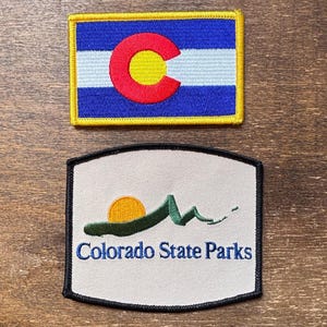May include: Two embroidered patches. The top patch is a rectangular Colorado state flag with a blue field, white stripes, and a red C with a yellow circle. The bottom patch reads "Colorado State Parks" with a mountain and sun design.