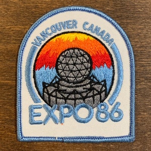May include: A white embroidered patch with blue edging. The patch features a stylized image of the Expo 86 logo, which includes a large, gray, geodesic dome with a red and orange sunset behind it. The text "Vancouver Canada Expo 86" is embroidered in blue around the dome.