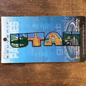 Pode incluir: Patch bordado com a palavra "UTAH" e imagens de marcos de Utah. O patch inclui imagens de uma cordilheira, uma formação rochosa e um edifício. O patch está em um fundo azul.