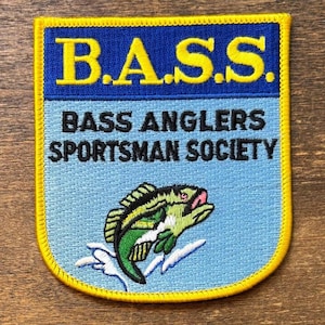 May include: A rectangular embroidered patch with a yellow border. The top section is dark blue with the yellow text B.A.S.S. The bottom section is light blue with the black text BASS ANGLERS SPORTSMAN SOCIETY and a green fish jumping out of water.