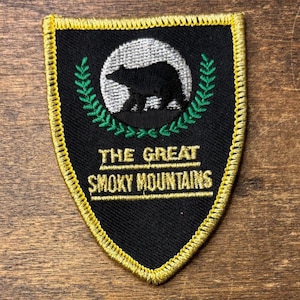 Könnte beinhalten: Ein schildförmiger, bestickter Aufnäher mit schwarzem Hintergrund und goldener Umrandung. Er zeigt ein silbernes kreisförmiges Emblem mit der Silhouette eines schwarzen Bären, umrahmt von grünen Lorbeerblättern. Der Text lautet THE GREAT SMOKY MOUNTAINS in Gold.