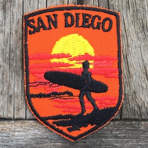 May include: A shield-shaped embroidered patch featuring an orange background with a yellow sun, red clouds, and a black silhouette of a person carrying a surfboard. The text SAN DIEGO is embroidered in black at the top of the patch.
