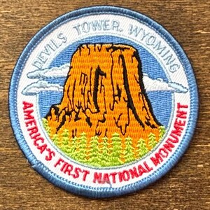 Puede incluir: Un parche bordado circular que presenta una representación de Devils Tower en Wyoming. El diseño incluye una formación rocosa de color naranja dorado, pinos verdes y un cielo azul con nubes blancas. El texto dice Devils Tower, Wyoming y America's First National Monument.