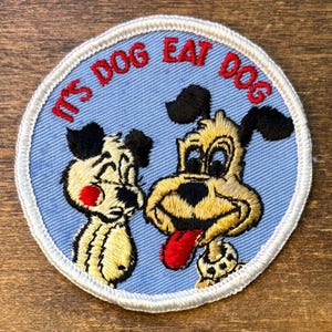 May include: Embroidered patch with a light blue background and a white border. The patch features two cartoon dogs with black ears and noses, and the text "IT'S DOG EAT DOG" in red. One dog has a red tongue.