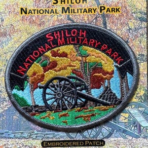 Pode incluir: Patch bordado com o texto "Shiloh National Military Park". O patch tem uma borda preta e mostra um canhão, árvores com folhagem de outono e o nome do parque em letras vermelhas e brancas. O patch tem formato oval.
