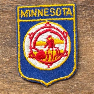 May include: A blue and yellow embroidered patch with the word "MINNESOTA" at the top. The patch features a circular emblem with a red and yellow design, including a person and landscape. The patch has a shield shape.