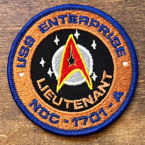Puede incluir: Parche bordado circular con borde azul y fondo naranja. Presenta la insignia USS Enterprise de Star Trek. El texto incluye "USS Enterprise Lieutenant NCC-1701-A". El emblema central es rojo, amarillo y negro.