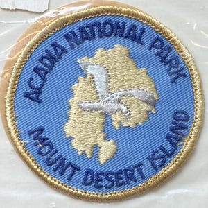 May include: A circular patch with a blue background and gold trim. The patch features the text "ACADIA NATIONAL PARK" and "MOUNT DESERT ISLAND" in blue. A light tan map of an island and a white seagull are in the center.