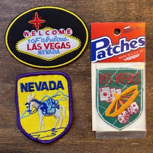 Könnte beinhalten: Vier gestickte Aufnäher mit Las Vegas- und Nevada-Motiven. Ein Aufnäher zeigt "Welcome to Fabulous Las Vegas Nevada". Ein weiterer zeigt einen Esel. Der dritte Aufnäher zeigt "Las Vegas" mit Würfeln und einem Rad. Der vierte Aufnäher ist verpackt und trägt die Aufschrift "Iron-On Patches".