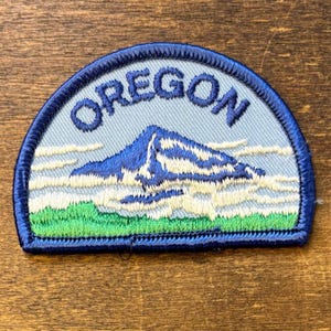 Könnte beinhalten: Ein halbkreisförmiger bestickter Aufnäher mit hellblauem Hintergrund und dem Wort OREGON in dunkelblauen Buchstaben. Das Design zeigt einen schneebedeckten Berggipfel in Weiß und Blau, umgeben von Wolken über einer grünen Grasfläche.